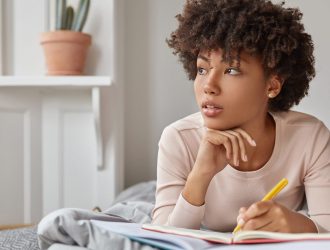 close-up-shot-pensive-dark-skinned-lady-with-afro-haircut-keeps-hand-chin-notes-text-notepad-with-yellow-pen-wears-casual-clothing-lies-comfortable-bed-focused-aside-rest-concept